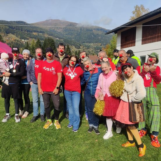 Eine Gruppe von Menschen, die rote Hemden und Clown-Make-up tragen, steht auf einem Rasen vor einem Gebäude mit Bergen im Hintergrund.