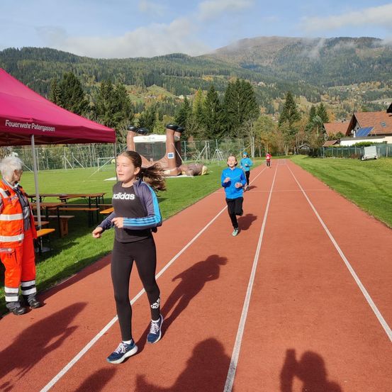 Ein junges Mädchen läuft auf einer Laufbahn mit Bergen im Hintergrund. Andere Läufer folgen. Eine ältere Person steht neben einem Zelt.