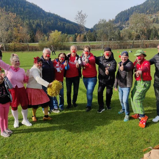 Eine Gruppe von Menschen mit roten Nasen und bunten Kostümen posiert für ein Foto auf einem Rasenfeld mit Bergen im Hintergrund.