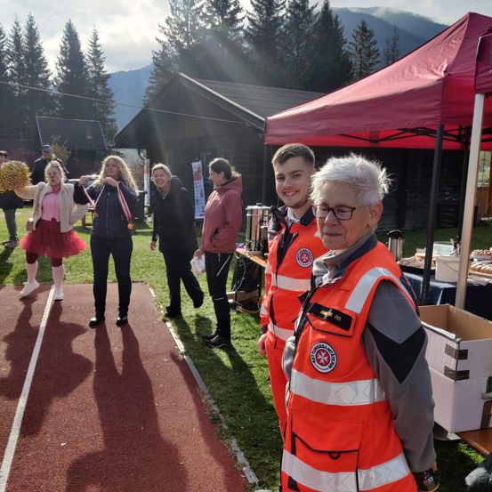 Mehrere Personen versammeln sich in der Nähe eines roten Zeltes auf einem Sportplatz. Eine Frau mit einer Pom-Pom steht in der Nähe der Laufbahn, während andere, einschließlich einer Frau in einem roten Tutu und einem Mann mit Brille, in der Nähe stehen.