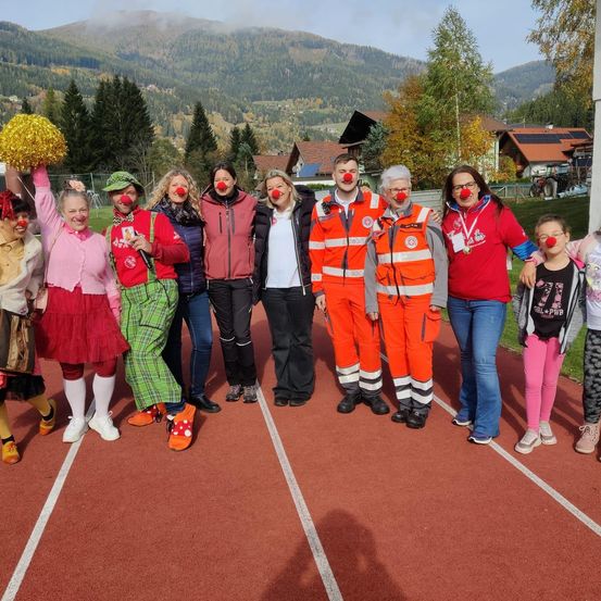 Eine Gruppe von Menschen in Clownkostümen mit roten Nasen posiert für ein Foto auf einer Laufbahn mit Bergen im Hintergrund.