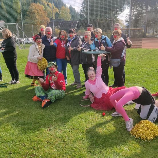 Eine Gruppe von Menschen in Clown-Kostümen posiert für ein Foto auf einem Rasenfeld. Einige sitzen, andere stehen. Der Hintergrund zeigt einen Zaun, Bäume und ein Gebäude.