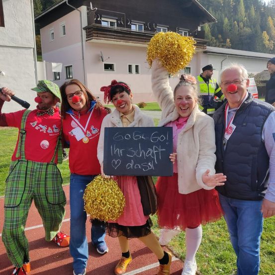 Eine Gruppe von Menschen in Clownkostümen posiert auf einer Laufbahn, ein Schild mit deutschem Text in der Hand. Einer hält eine Pom-Pom, der andere ein Mikrofon. Hinter ihnen sind Gebäude und ein Mann in einem gelben Weste zu sehen.