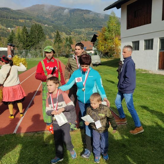 Eine Gruppe von Menschen steht auf einem Grasfeld mit Bergen im Hintergrund. Zwei Jungen und eine Frau halten Auszeichnungen. Ein Mann mit roter Nase und eine Frau in einer grünen Mütze stehen in ihrer Nähe. Ein Mädchen mit einer Pom-Pom und ein Mann in einer blauen Jacke sind in der Nähe.