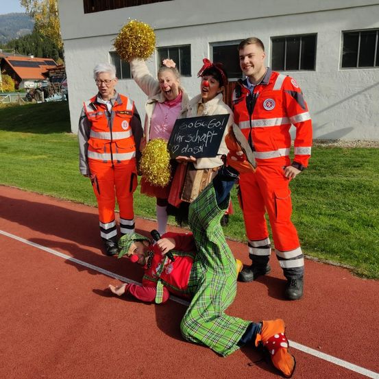 Eine Gruppe von Menschen in roten Sicherheitsanzügen steht auf einer Laufbahn, einer kniet mit Clownsnase. Hinter ihnen hält eine Frau ein Schild und Pom-Poms.