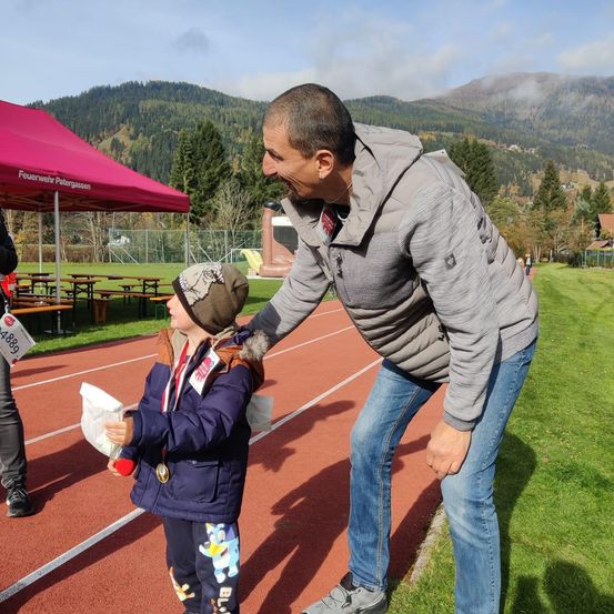 Ein Mann lacht mit einem kleinen Kind, das eine Medaille und einen Plastikbeutel hält, auf einer Laufbahn. Im Hintergrund befindet sich ein rotes Zelt, ein Zaun und ein Berg.