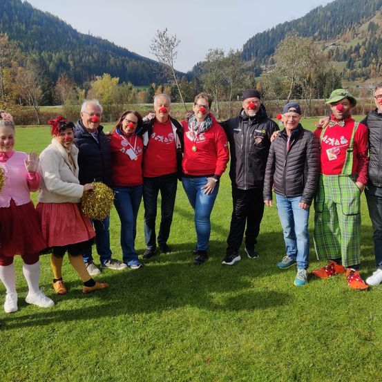 Eine Gruppe von Menschen, die in Rot gekleidet und mit Clown-Make-up und Accessoires geschminkt sind, posiert für ein Foto auf einer Wiese mit Bergen im Hintergrund.