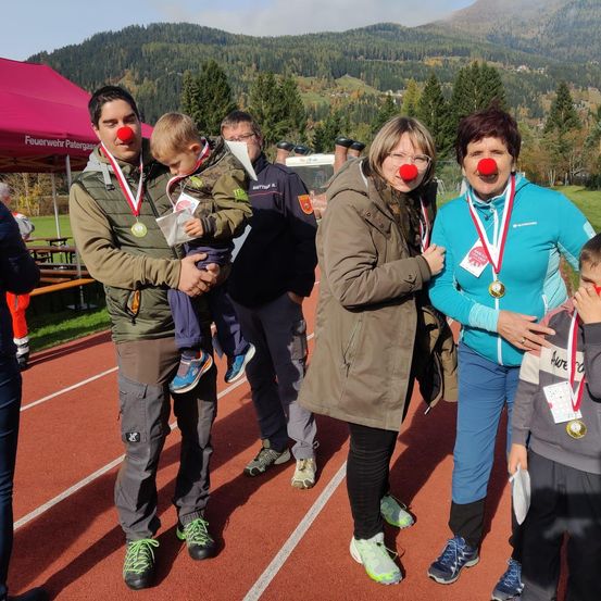Mehrere Menschen stehen auf einer Laufbahn, zwei Frauen mit Clown-Nasen. Ein Mann hält ein Kind. Dahinter steht ein Mann in einem schwarzen Uniform. Bäume und Berge sind im Hintergrund.