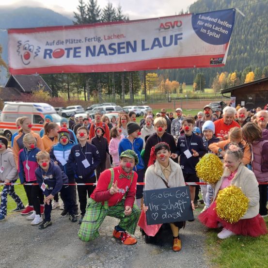 Eine Gruppe von Menschen in Clownkostümen versammelt sich auf einem Feld mit einem Banner über ihnen, das 'Rote Nasen Lauf' liest. Sie halten Schilder und jubeln.