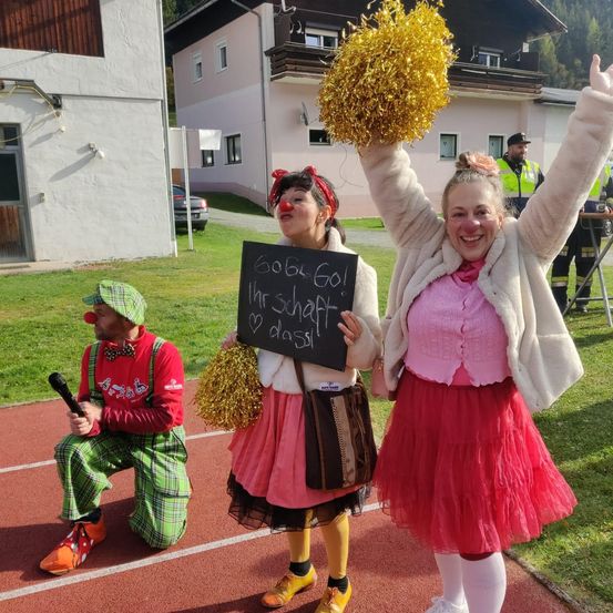 Drei Personen posieren auf einer Laufbahn. Zwei sind in rosa Röcken gekleidet, halten Pom-Poms und lächeln, während die dritte ein Clown vor ihnen kniet.