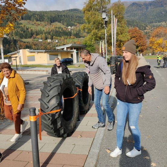 Vier Personen stehen neben großen Reifen auf einer Stadtstraße. Ein Mann betrachtet die Reifen, während ein anderer Mann lächelt. Zwei Frauen stehen in der Nähe, eine mit den Händen in den Taschen. Hinter ihnen ist ein Gebäude mit einem geparkten Auto sichtbar. Bäume und Berge sind im Hintergrund zu sehen.