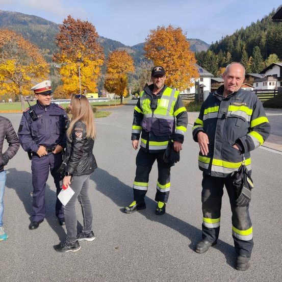Zwei Polizisten und zwei Feuerwehrleute stehen mit einer Frau in einer Jacke draußen. Sie befinden sich auf einem Parkplatz mit Bäumen und Bergen im Hintergrund.