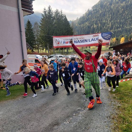 Eine Gruppe von Kindern läuft bei einem Rennen. Ein Mann in einem roten Kostüm mit Clownschuhen feuert sie an. Über ihnen hängt ein Banner mit den Worten "Rot Nasen Lauf".