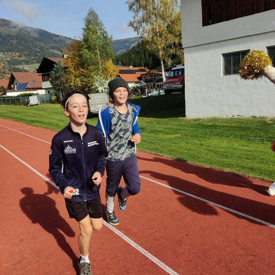 Zwei Jungen laufen auf einer Laufbahn mit Bergen im Hintergrund. Sie tragen Sportkleidung und Stirnbänder. Einer hält einen roten Ball.