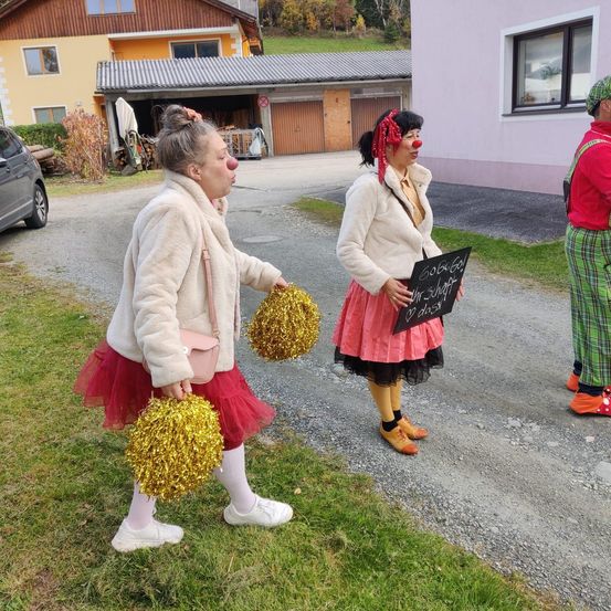 Zwei als Clowns verkleidete Frauen gehen mit einem Schild auf einer Auffahrt, eine hält Pom-Poms und die andere hat eine rote Bandana und eine rote Nase.