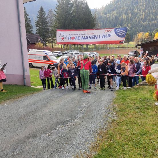 Eine Menschenmenge, einige in Clownkostümen, steht in einer Reihe an einer roten Absperrung. Ein Banner oben besagt 'Rote Nasen Lauf'. Dahinter ist ein Rettungsfahrzeug geparkt.