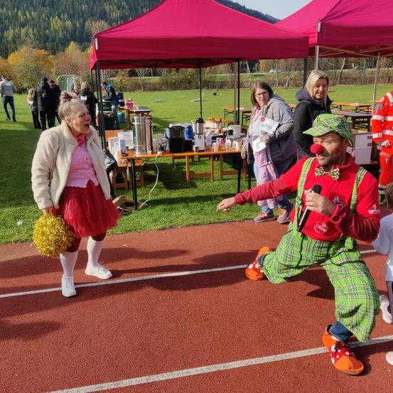 Ein Mann in Clown-Kleidung kniet auf einer Laufbahn, während eine Frau in einer Cheerleader-Uniform Pom-Poms hinter ihm hält. Im Hintergrund stehen rote Zelte und Picknicktische.