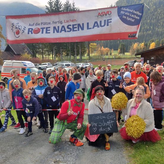 Eine Menschenmenge mit roten Nasen posiert für ein Foto. Ein Banner mit Rote Nasen Lauf und ASVO steht im Vordergrund. Sie befinden sich vor einem Feld mit einem Berg im Hintergrund.