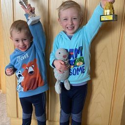 Zwei junge Jungen stehen nebeneinander. Sie tragen beide blaue Hoodies und Hosen. Sie halten Pokale und lächeln. Ein Junge hält ein Kuscheltier.