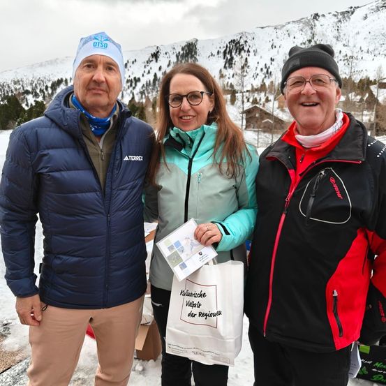 Drei Personen, in Winterkleidung gekleidet, posieren für ein Foto vor einem verschneiten Berg und halten einen Beutel mit der Aufschrift 'Kulinarische Vielfalt der Region'.