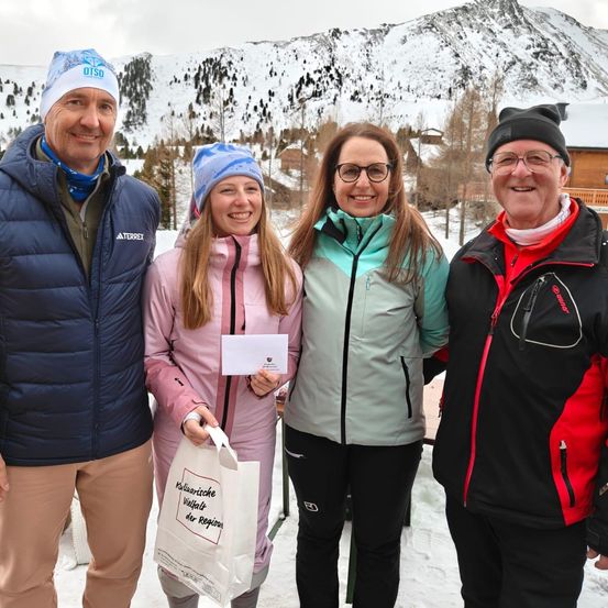 Vier Personen stehen im Schnee mit einer Bergkette im Hintergrund. Sie alle lächeln. Die Frau links hält einen Beutel. Die Frau in der Mitte hält ein Stück Papier. Der Mann rechts trägt eine Mütze.
