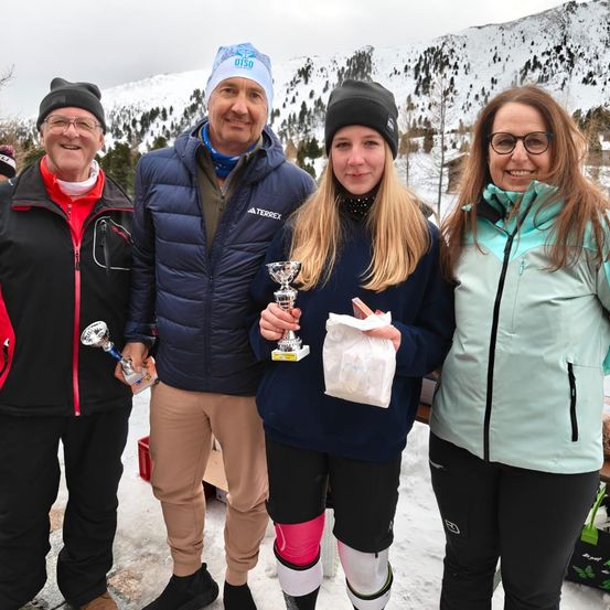 Vier Menschen stehen im Schnee mit Bergen im Hintergrund. Zwei Männer und zwei Frauen, alle lächeln. Zwei von ihnen halten Trophäen und eine Frau hält einen Beutel.