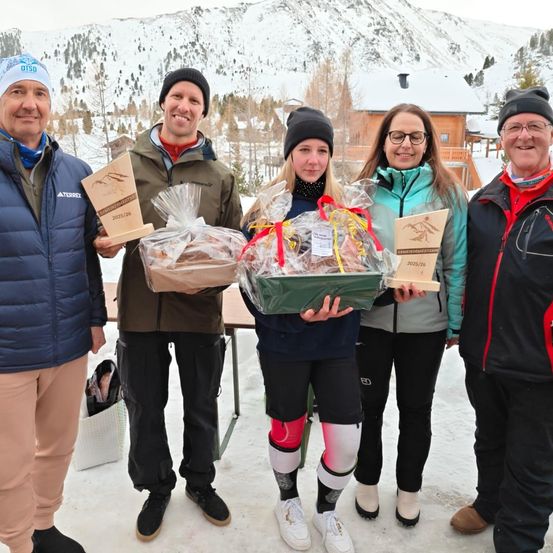 Fünf Menschen stehen auf einer verschneiten Oberfläche und halten eingewickelte Geschenke und Trophäen. Sie sind alle in Winterkleidung gekleidet. Die Frau in der Mitte hält einen Korb. Hinter ihnen steht ein Holzhaus mit Schnee auf dem Dach.