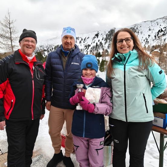 Eine vierköpfige Familie steht im Schnee und lächelt. Das Kind hält einen Pokal. Sie tragen Winterkleidung. Schneebedeckte Berge sind im Hintergrund.