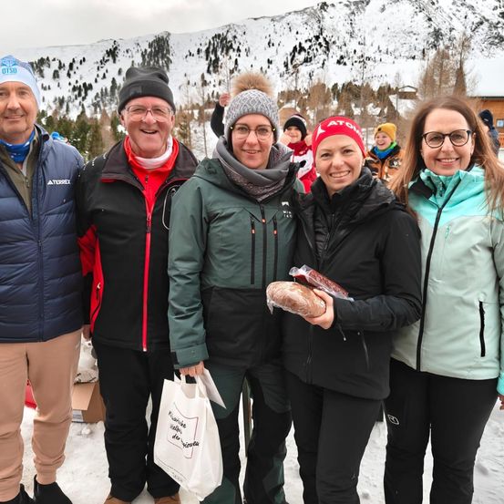 Eine Gruppe Erwachsener in Winterkleidung steht im Schnee und lächelt für ein Foto. Eine Person hält ein Sandwich. Schneebedeckte Berge sind im Hintergrund.