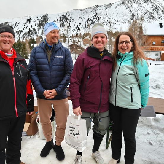 Vier Menschen, für kaltes Wetter gekleidet, stehen im Schnee und lächeln. Hinter ihnen ist ein schneebedeckter Berg und ein Holzhaus. Ein Mann hält einen Beutel.