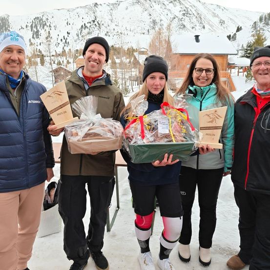 Fünf Menschen posieren lächelnd im Schnee für ein Foto. Sie halten verpackte Geschenke. Hinter ihnen ist ein Haus mit einem Schornstein zu sehen, und im Hintergrund sind mit Schnee bedeckte Berge.