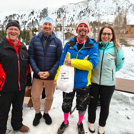 Vier Personen stehen in einem verschneiten Bereich und lächeln, mit schneebedeckten Bergen im Hintergrund. Sie tragen Winterkleidung. Der Mann auf der linken Seite hält eine Tasche.