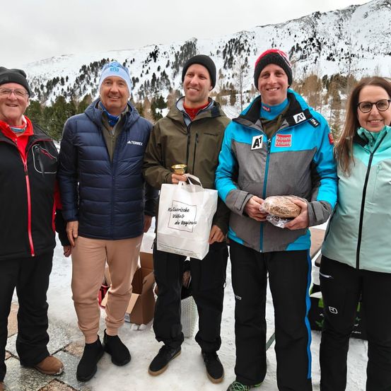 Fünf Personen stehen gemeinsam in einem verschneiten Berggebiet. Sie alle lächeln. Drei von ihnen halten Tassen, und eine hält einen Papierbeutel. Der Berg ist schneebedeckt.