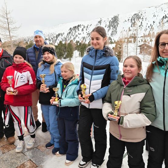 Eine Gruppe junger Mädchen und Erwachsener steht im Schnee und hält Pokale. Sie alle lächeln. Hinter ihnen sind verschneite Berge.