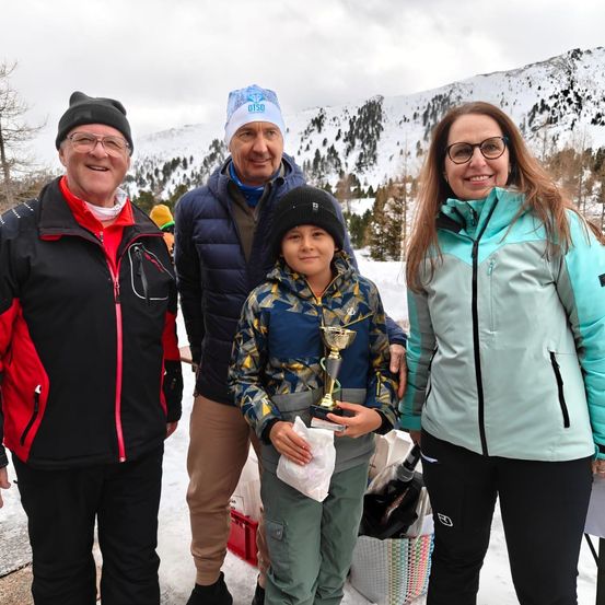 Vier Menschen stehen im Schnee und lächeln. Ein Junge hält einen Pokal. Im Hintergrund sind verschneite Berge und Bäume zu sehen.