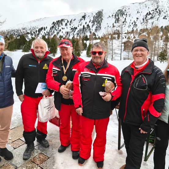 Fünf Männer stehen im Schnee und tragen Winterkleidung. Drei von ihnen tragen rote Hosen und schwarze Jacken. Zwei Männer tragen Medaillen. Der Hintergrund besteht aus verschneiten Bergen und Häusern.
