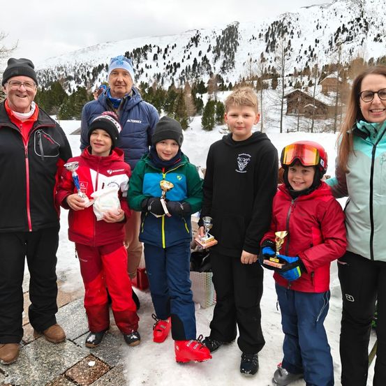 Eine Familie mit Kindern in Winterkleidung steht lächelnd auf einem verschneiten Berg. Jedes Kind hält eine Trophäe und Erwachsene halten Tassen. Schneebedeckte Berge und Bäume sind im Hintergrund.