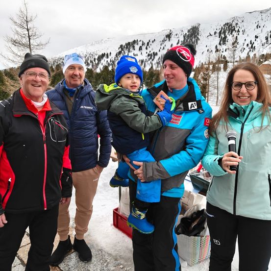 Fünf Personen stehen im Schnee, zwei halten ein Kind. Der Mann hält ein Mikrofon, die Frau trägt eine Brille. Sie lächeln, das Kind trägt eine Mütze. Hinter ihnen sind schneebedeckte Berge und Bäume.