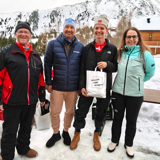 Vier Personen posieren für ein Foto im Schnee, mit Taschen. Dahinter ein Berg mit einem Haus in der Ferne.