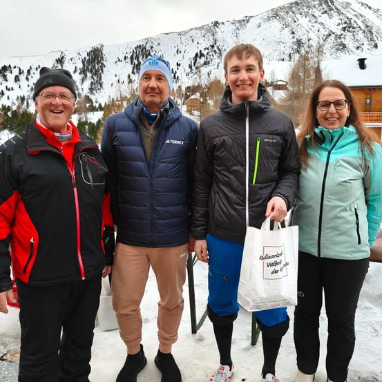 Vier Personen stehen im Schnee und tragen Winterkleidung. Der Mann auf der linken Seite trägt eine rot-schwarze Jacke. Der Mann neben ihm trägt eine blaue Jacke mit blauer Kappe. Der Mann rechts trägt eine schwarze Jacke und blaue Hose. Die Frau rechts trägt eine grün-schwarze Jacke.