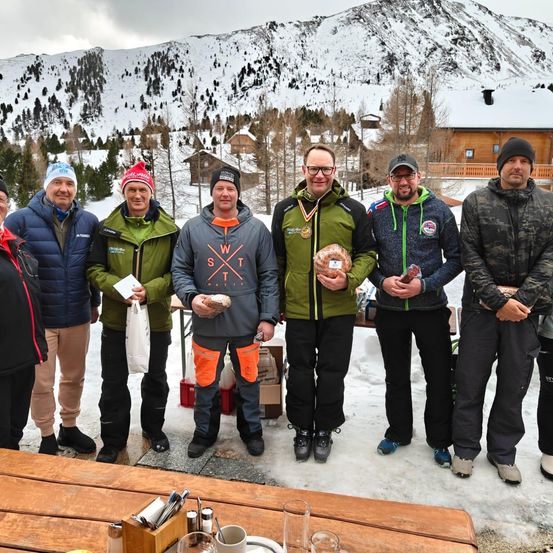 Eine Gruppe von Männern in Winterkleidung steht auf einem verschneiten Berg, lächelt und posiert für ein Foto. Sie halten Brot und einige haben Medaillen. Ein Holztisch mit Tassen und Gewürzen steht davor.