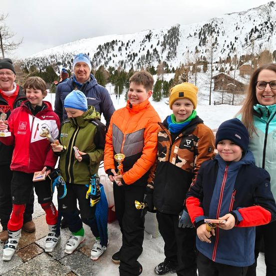 Eine Gruppe von Kindern und Erwachsenen steht im Schnee, lächelt und hält Pokale in der Hand, mit einem verschneiten Berg im Hintergrund.