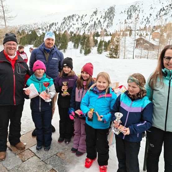 Mehrere Menschen, darunter Erwachsene und Kinder, stehen im Schnee, lächeln und halten Trophäen. Sie tragen Winterkleidung. Ein Haus ist in der Ferne sichtbar, und das Gebiet ist schneebedeckt.
