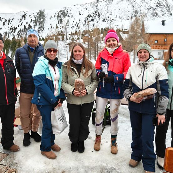 Eine Gruppe von Menschen in Winterkleidung, die lächelnd und für ein Foto im Schnee posieren, mit Bergen und Häusern im Hintergrund.