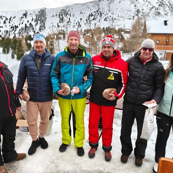 Eine Gruppe von Männern in Winterkleidung posiert für ein Foto im Schnee mit Brot in den Händen. Dahinter befindet sich ein schneebedeckter Berg und ein Chalet.