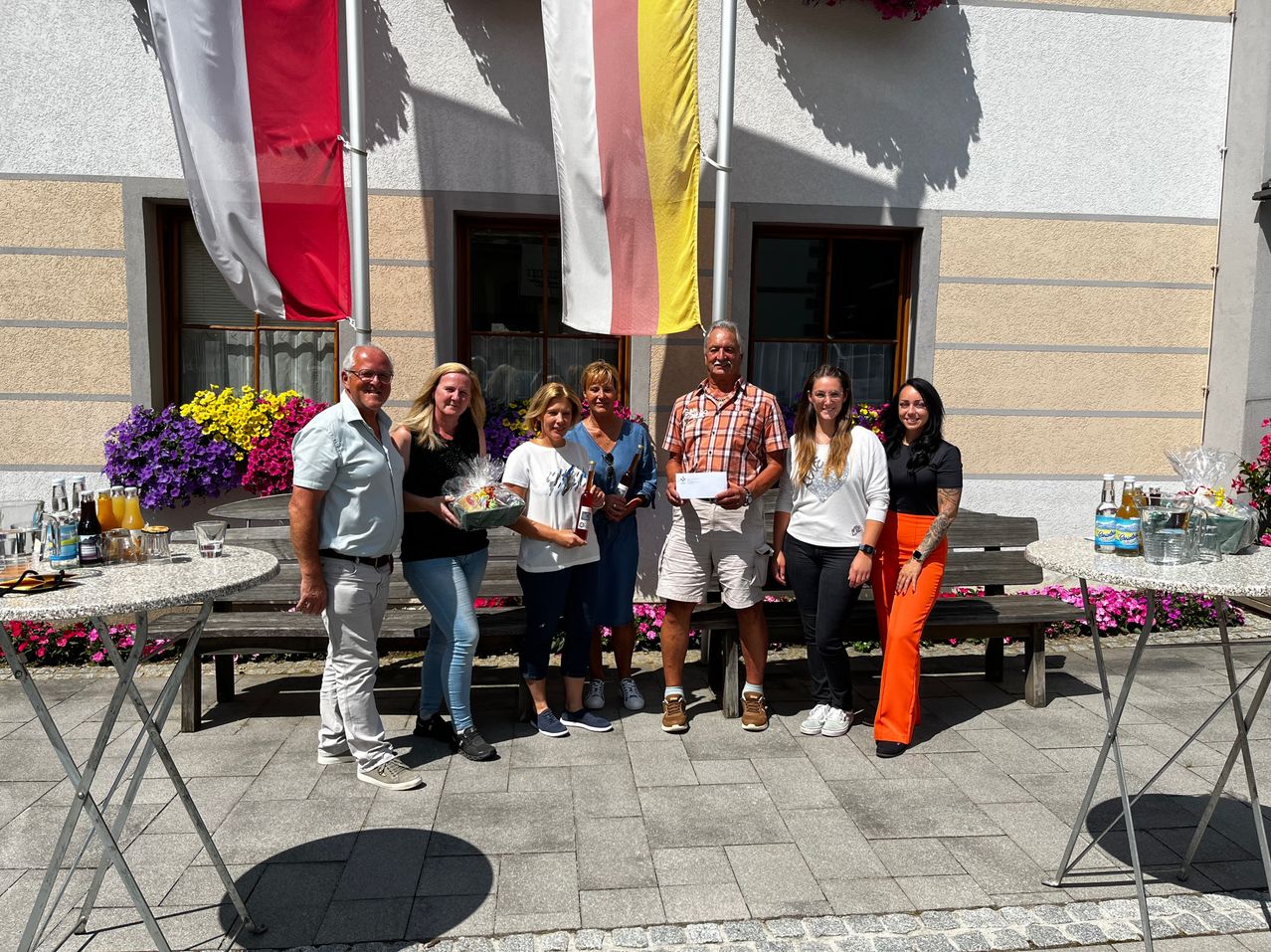Sieben Personen posieren für ein Foto vor einem Gebäude mit Fahnen. Eine Frau hält einen Korb und ein Zertifikat, und ein Mann hält eine Flasche. Dahinter stehen Bänke und Blumen.
