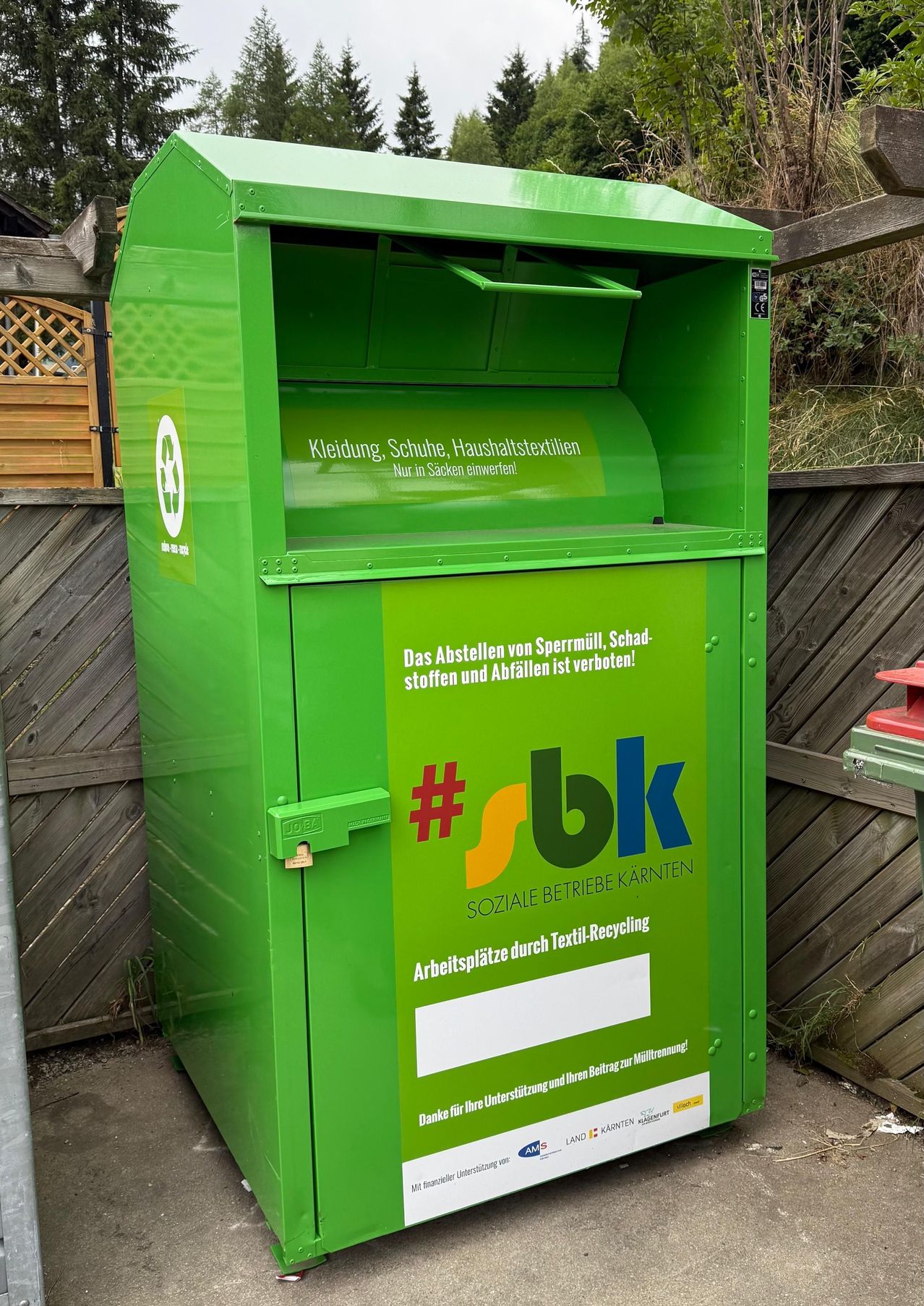 Ein grüner Recyclingbehälter mit weißem Deckel hat ein Etikett mit dem Recycling-Symbol. Der Text auf dem Behälter lautet: #bk Soziale Betriebe Karnten, Arbeitsplatze durch Textil-Recycling.