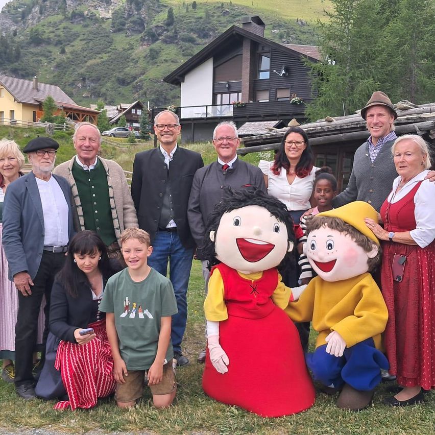 Eine Gruppe von Erwachsenen und Kindern posiert für ein Foto vor einem Haus mit einem Berg im Hintergrund. Sie lächeln und einige tragen Kostüme.