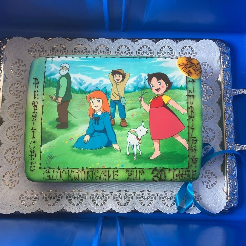 Ein Kuchen ist in einer blauen Schachtel mit einem Spitzendeckchen ausgestellt. Der Kuchen hat ein Cartoon-Bild von drei Personen und einem Ziegenbock. Eine blaue Schleife ist um den Kuchen gebunden. Der Kuchen hat die Worte 'Gluckwunsche zum 30th' darauf geschrieben.