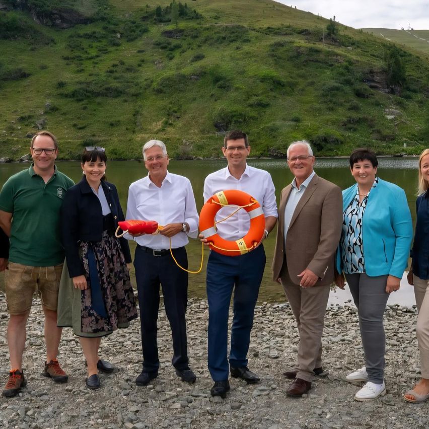 Eine Gruppe von Personen steht am Ufer eines Flusses mit einem Berg im Hintergrund. Sie alle lächeln und posieren für ein Foto. Ein Mann hält eine Rettungsboje.